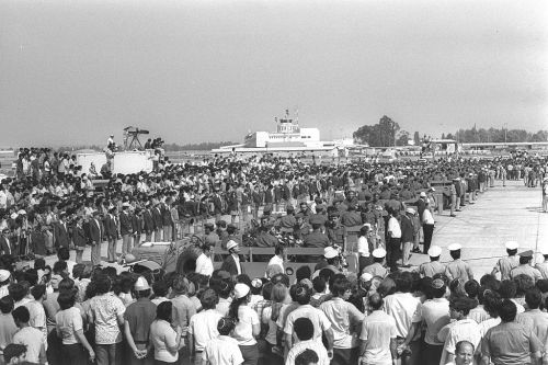 A large crowd gathers at an airport, with military personnel and vehicles in the foreground, under a clear sky.