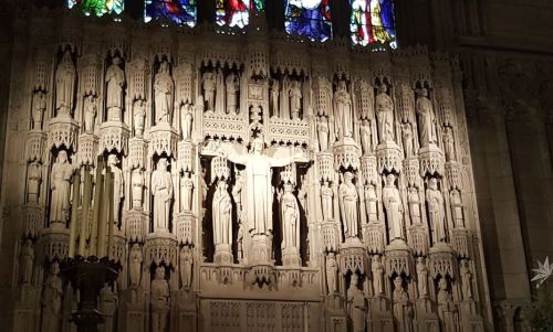 Intricate stone wall with statues and stained glass windows, featuring a central figure on a cross.