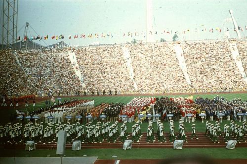 A large crowd at a stadium during a colorful opening ceremony with flags and performers in coordinated outfits.