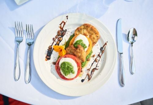 A plate of colorful layered vegetables topped with pesto and a flower garnish, served with utensils on a white tablecloth.