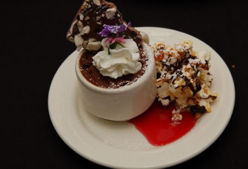 A dessert plate featuring a chocolate mousse topped with whipped cream, a chocolate shard, and popcorn drizzled with chocolate.