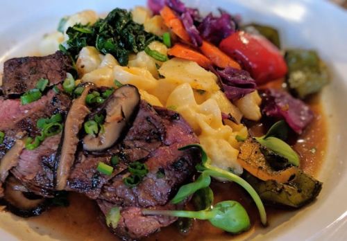 A plate of sliced steak with mushrooms, served alongside pasta and a colorful mix of sautéed vegetables.