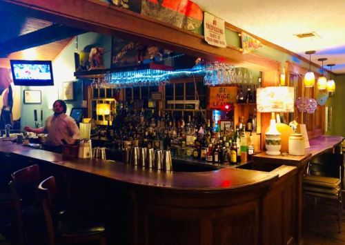 A cozy bar with a wooden counter, shelves of liquor, and warm lighting, featuring a bartender and a TV in the background.