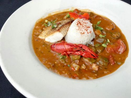 A bowl of gumbo with fish, rice, crawfish, and vegetables, garnished with green onions and spices.