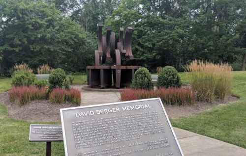 A bronze sculpture titled "David Berger Memorial" surrounded by greenery and a plaque detailing its significance.