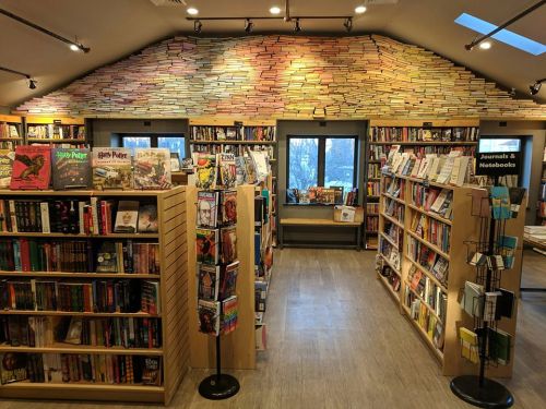 Cozy bookstore interior with wooden shelves filled with books, a unique wall of stacked books, and large windows.