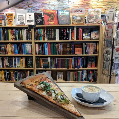 A slice of pizza on a wooden stand with a bowl of soup, set against a colorful bookshelf filled with books.