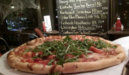 A large pizza topped with fresh tomatoes and arugula, placed on a table with a chalkboard menu in the background.