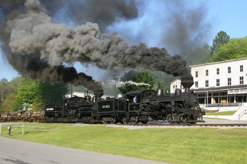 A vintage steam locomotive emits black smoke while traveling along a track near a historic building.
