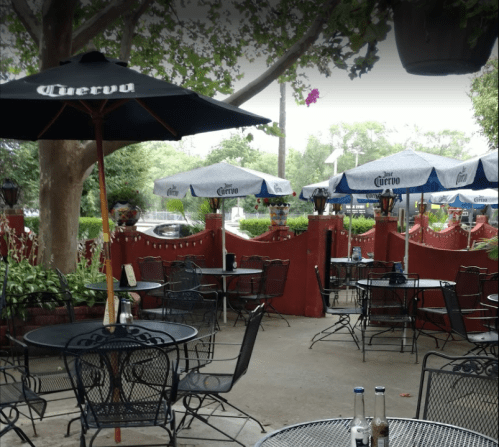 Outdoor dining area with black metal tables and chairs, shaded by umbrellas, surrounded by greenery and a red wall.