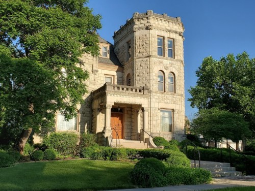 A historic stone mansion with a turret, surrounded by lush greenery and a clear blue sky.