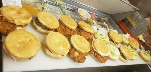 A variety of assembled burgers and chicken sandwiches on a countertop, with toppings and condiments in the background.