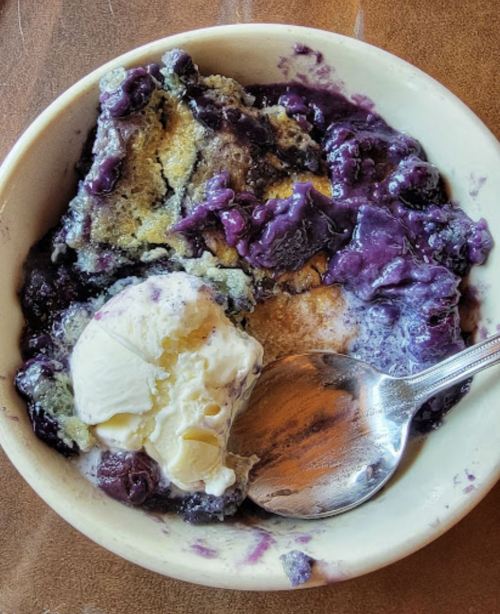 A bowl of blueberry cobbler topped with a scoop of vanilla ice cream and a spoon resting beside it.