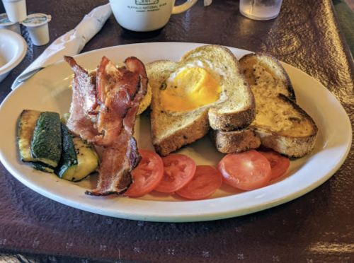A plate of breakfast featuring bacon, egg in toast, grilled zucchini, and sliced tomatoes.