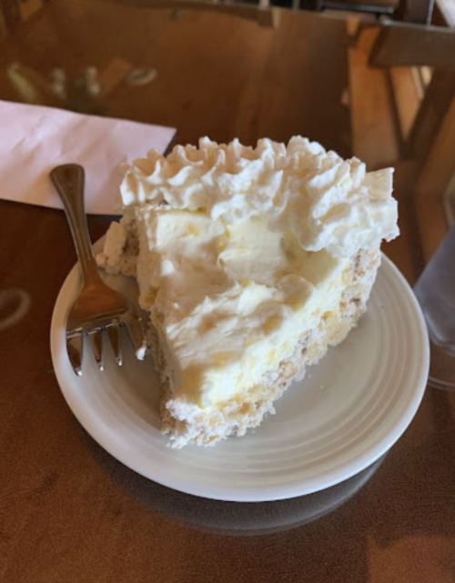 A slice of creamy cake topped with whipped cream on a white plate, accompanied by a fork.