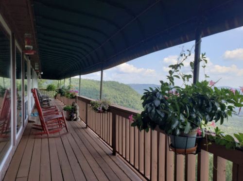 A scenic porch with rocking chairs and hanging plants, overlooking lush green mountains under a blue sky.