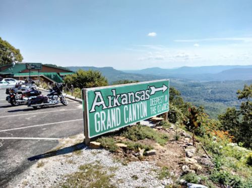 Sign for Arkansas Grand Canyon with motorcycles parked nearby, overlooking a scenic mountainous landscape.