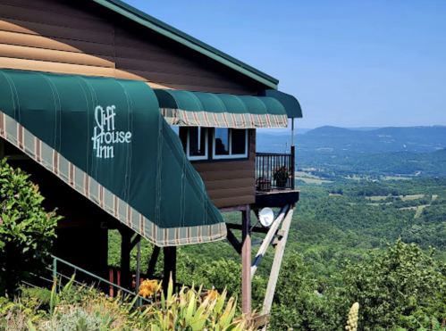 A mountain inn with a green awning, overlooking a scenic valley and distant hills under a clear blue sky.