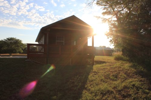A small wooden house with a porch, illuminated by sunlight, surrounded by grass and trees under a blue sky.