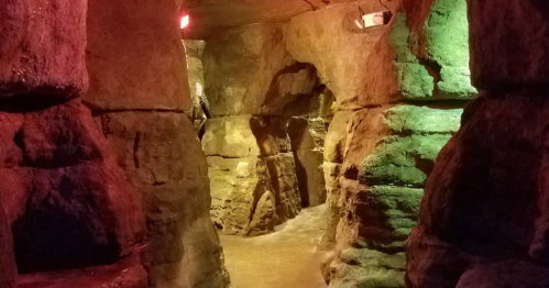 A dimly lit stone corridor with colorful lights, featuring rough, textured walls and a winding path.
