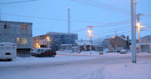 Snowy street scene with buildings, power lines, and streetlights under a blue sky at dusk.