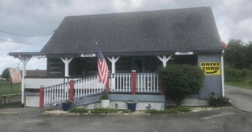 A small, rustic building with a drive-thru sign, American flag, and colorful flower pots on the porch.