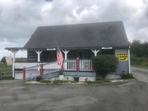 A small, rustic building with a drive-thru sign and an American flag, surrounded by greenery and cloudy skies.
