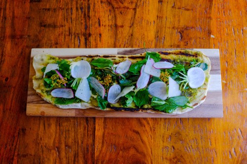A flatbread topped with greens, radish slices, and herbs, served on a wooden board against a rustic table.