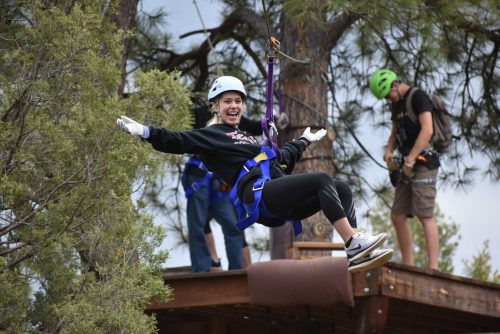 A person joyfully zip-lining from a platform, wearing a helmet and harness, with trees and another person in the background.