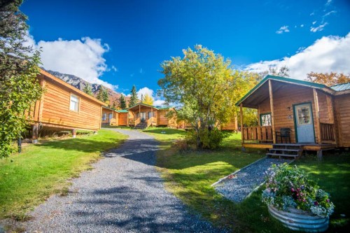 A scenic view of wooden cabins along a gravel path, surrounded by green grass and trees under a blue sky.