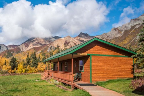 A rustic wooden cabin surrounded by colorful autumn foliage and mountains under a blue sky with fluffy clouds.