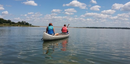 Two people in a canoe paddling on a calm lake under a blue sky with fluffy clouds.
