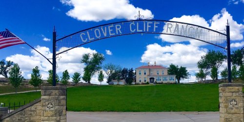 Archway reading "Clover Cliff Ranch" with a historic building and green landscape under a blue sky with clouds.