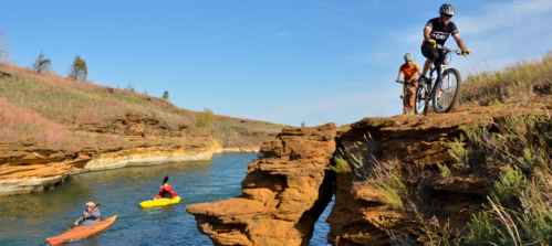 A cyclist navigates rocky terrain while two kayakers paddle in a serene river surrounded by grassy hills.