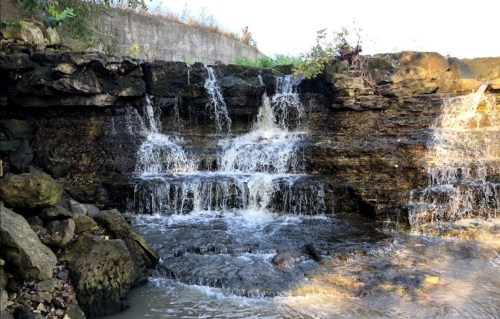 A small waterfall cascading over rocky terrain into a calm body of water, surrounded by greenery.