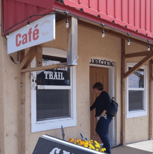 A woman approaches a café entrance with a welcoming sign and colorful flowers in front.