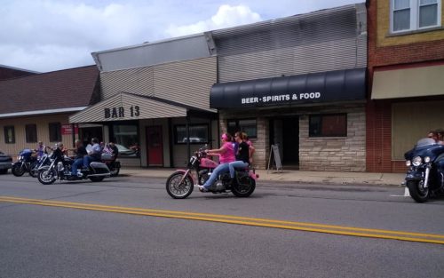A group of motorcycles parked outside a bar, with riders socializing on the street in a small town setting.