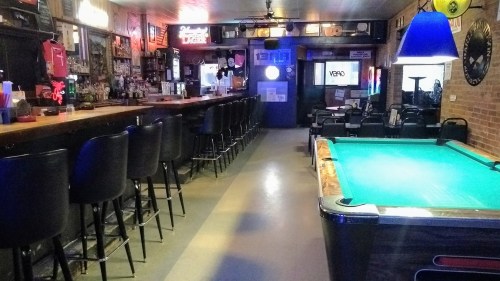 A dimly lit bar interior featuring a long counter, bar stools, and a pool table in the foreground.