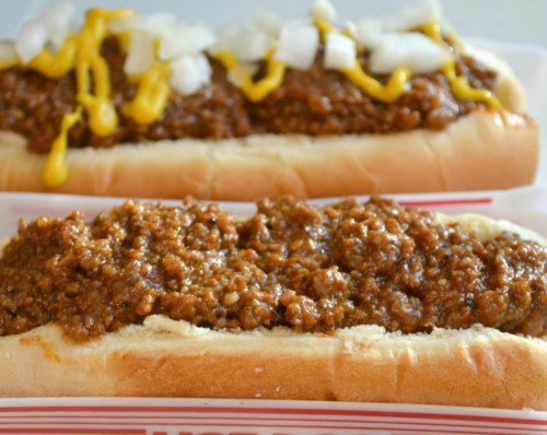 Two hot dogs topped with chili, mustard, and diced onions, served in red and white paper trays.