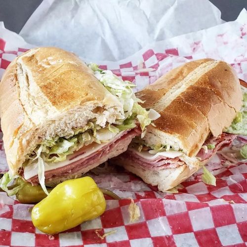 A freshly cut sub sandwich filled with layers of meat, lettuce, and cheese, served with pickled peppers on a checkered paper.