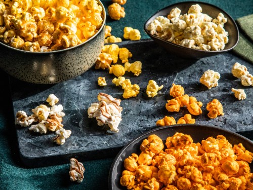 A variety of popcorn in bowls and scattered on a dark surface, featuring yellow, white, and orange colors.
