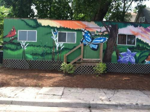 Colorful mural on a green trailer featuring a butterfly, flowers, and a cardinal, surrounded by greenery and mulch.