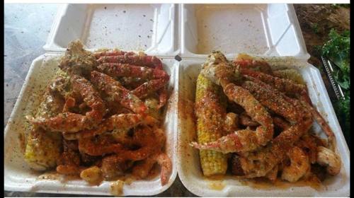 Two takeout containers filled with seasoned seafood, including crab legs, shrimp, and corn on the cob.