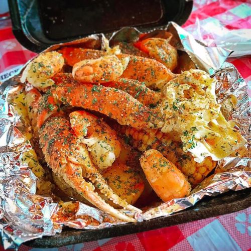 A takeout container filled with seasoned crab legs, shrimp, and corn, garnished with herbs on a red checkered tablecloth.