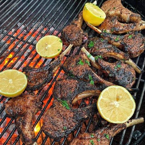 Grilled lamb chops with a charred crust, garnished with lemon slices, sizzling on a barbecue grill.