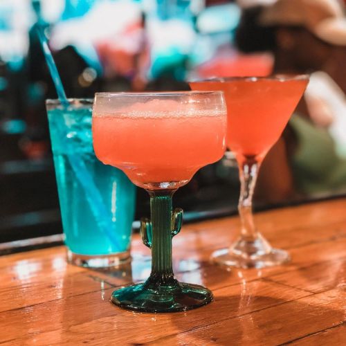 Three colorful cocktails on a wooden bar: a blue drink, a pink margarita, and a pink martini.