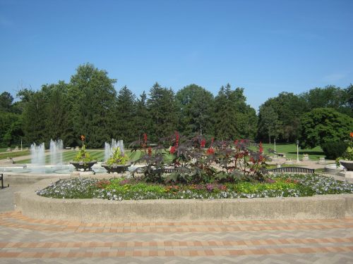 A vibrant garden with colorful flowers and fountains, surrounded by lush green trees under a clear blue sky.