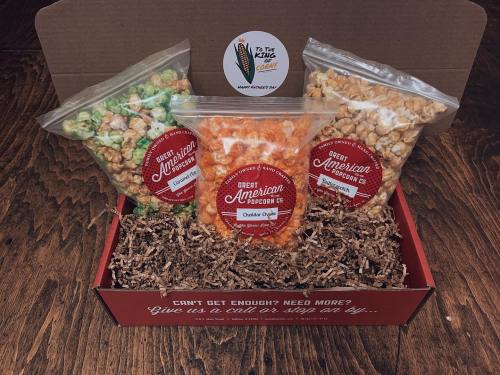 A gift box containing three bags of flavored popcorn: green, orange, and yellow, on a bed of shredded paper.