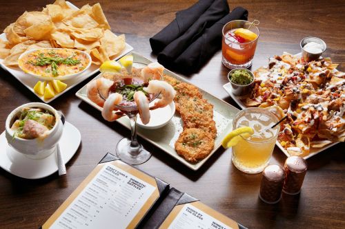 A table spread with nachos, shrimp cocktail, seafood dish, and drinks, surrounded by chips and garnishes.