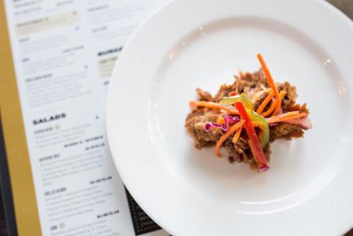 A small serving of pulled meat topped with colorful shredded vegetables on a white plate, with a menu in the background.